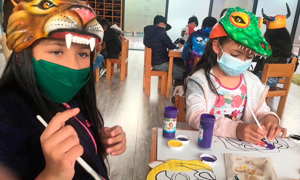 Foto de dos niñas estudiantes mostrando sus trabajos, tienen gorros en forma de animal en la cabeza