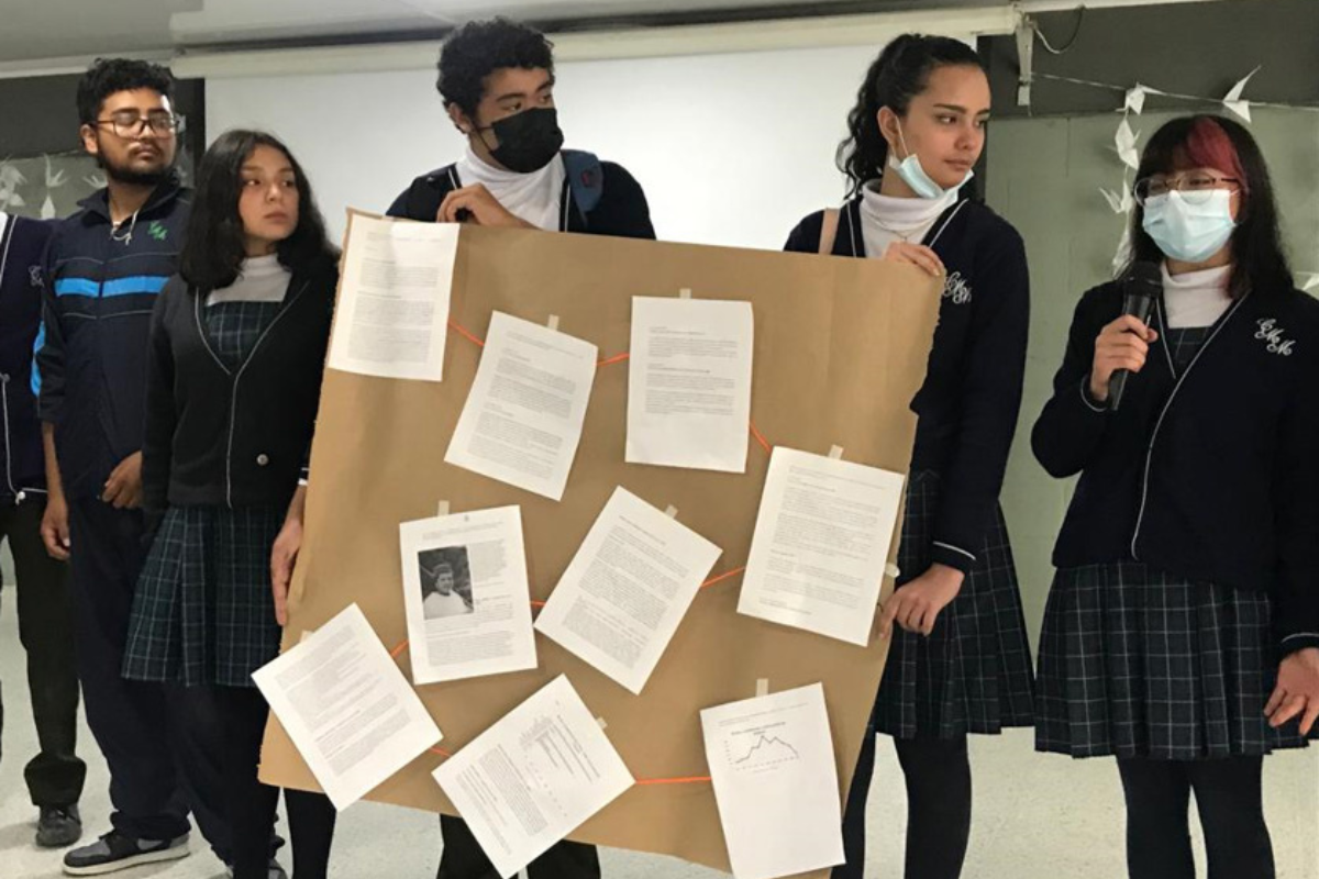 Foto de un grupo de 5 estudiantes exponiendo una cartelera con varias hojas pegadas allí
