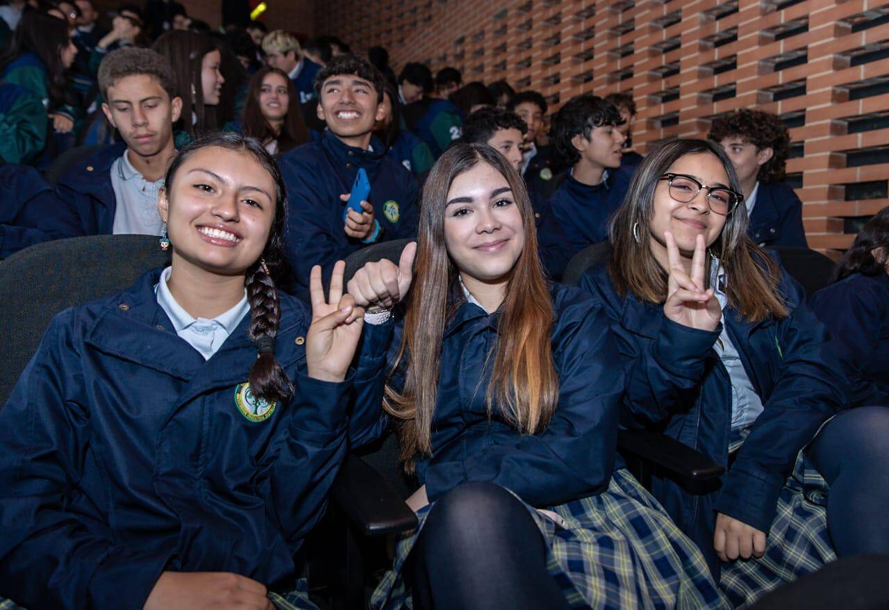 Foto de 3 estudiantes alegres en un auditorio con más compañeros atrás